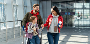 A New Zealand family with school-aged child arriving home at the airport with luggage, warm summer light through the windows suggesting the start of a new school year.